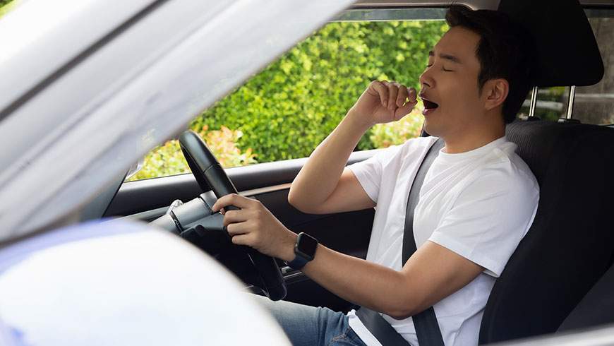 Yawning, a tired young man sits in his car after a long day of work, increasing his risk of an accident