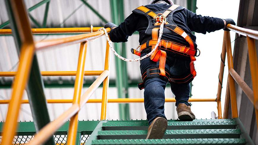 A construction worker climbs high steps, wearing a safety harness and safety line
