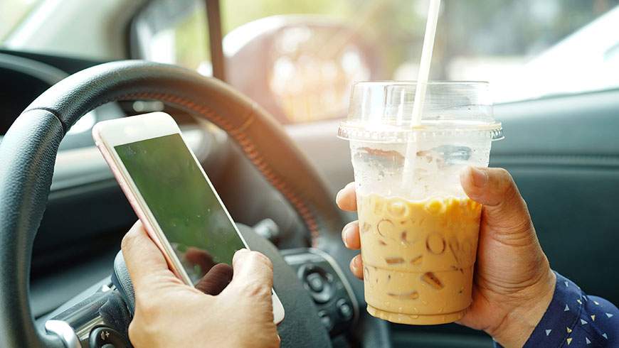 A woman distracted in her car holds her phone and an iced coffee while behind the wheel
